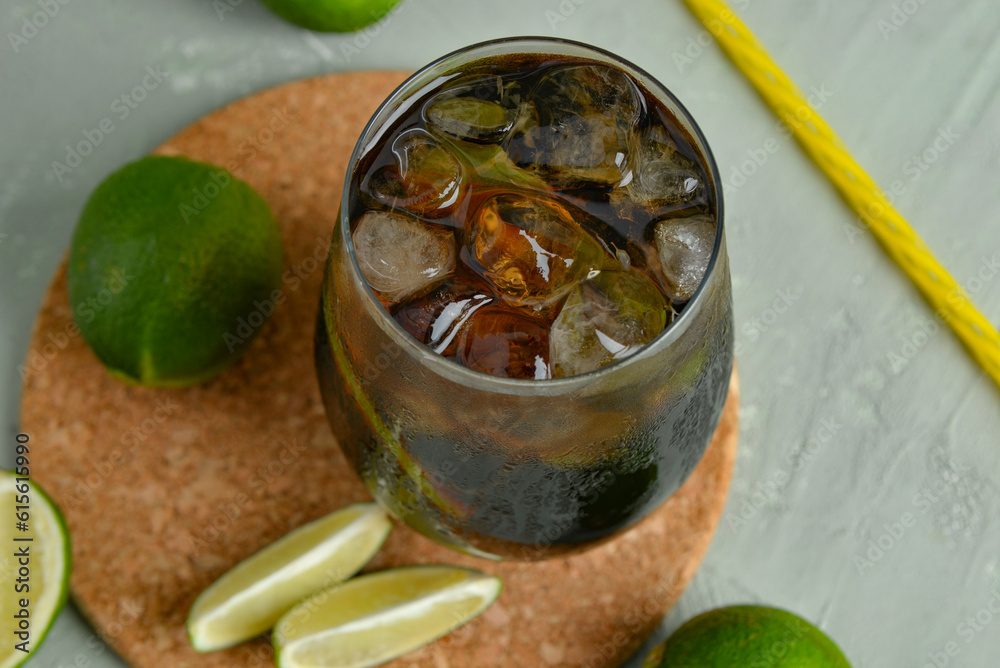 Glass of cold Cuba Libre cocktail and limes on grey background