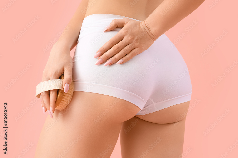 Young woman massaging her leg with anti-cellulite brush on pink background, back view