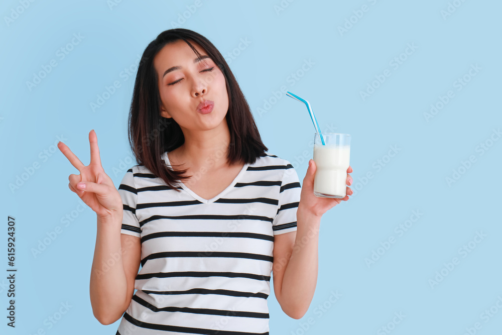 Beautiful Asian woman with glass of milk showing victory gesture on light blue background