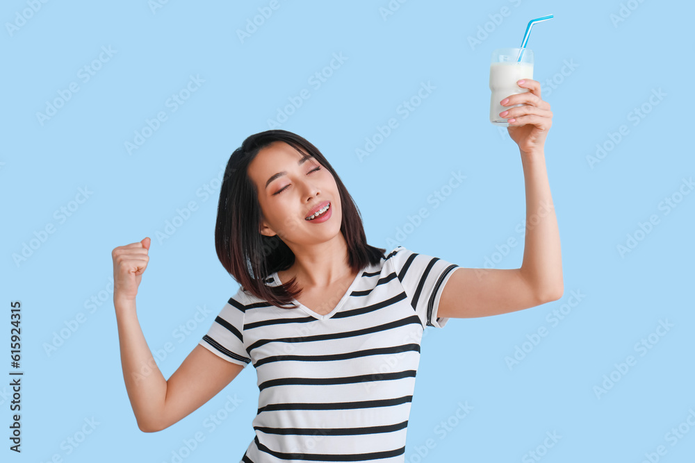 Happy Asian woman with glass of milk on light blue background