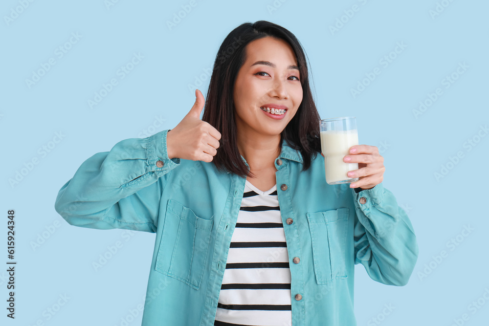 Beautiful Asian woman with glass of milk showing thumb-up on light blue background