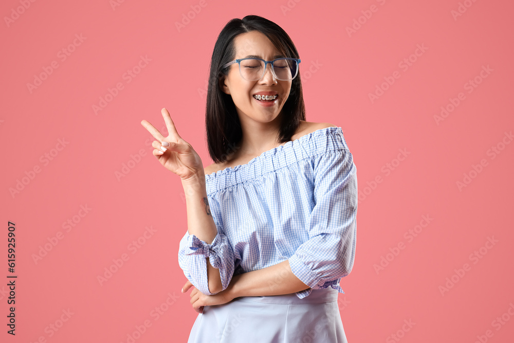 Beautiful Asian woman in stylish eyeglasses showing victory gesture on pink background