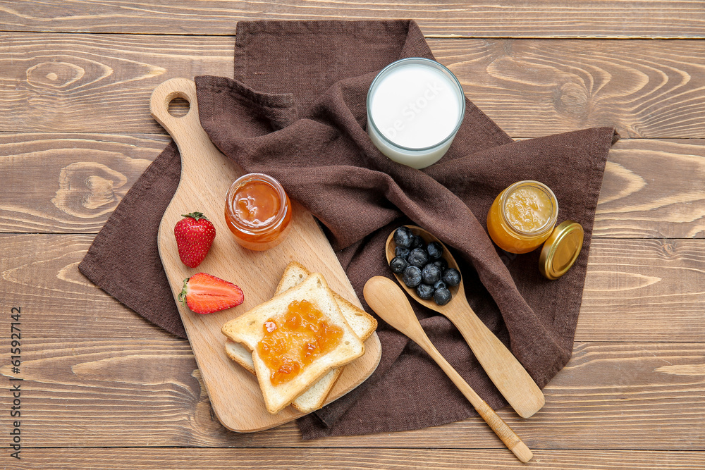 Wooden board with tasty toasts, jars of different jams and milk on brown wooden background