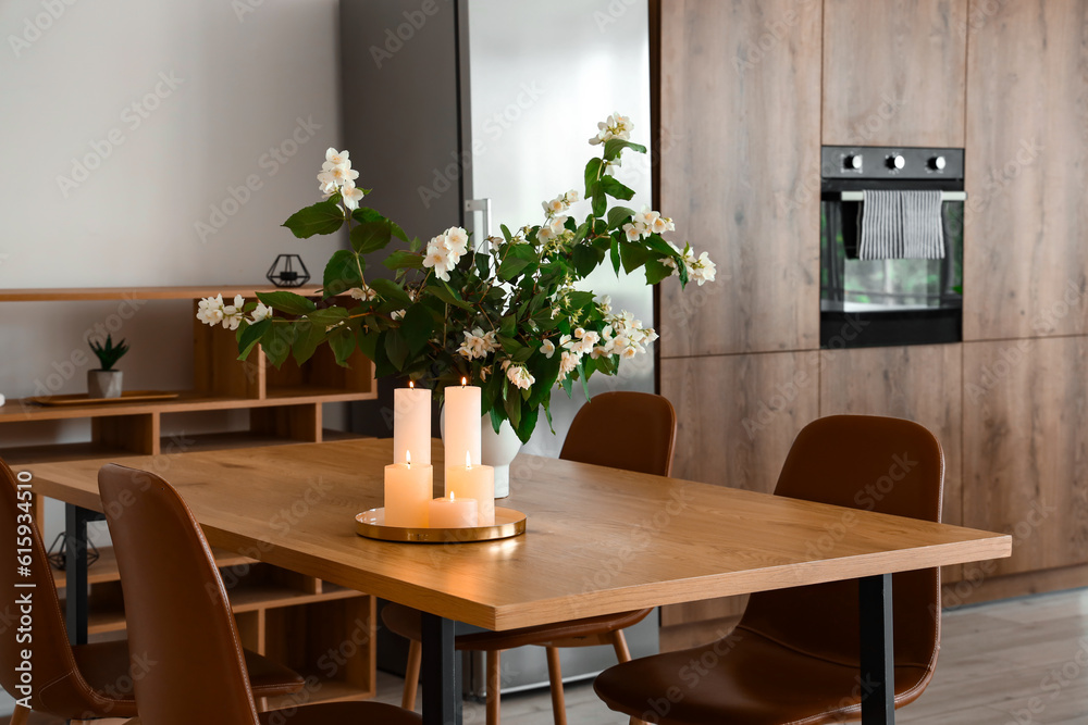 Vase with blooming jasmine flowers and burning candles on wooden table in modern kitchen