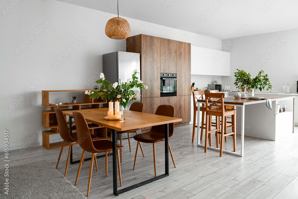 Vase with blooming jasmine flowers and burning candles on wooden table in modern kitchen