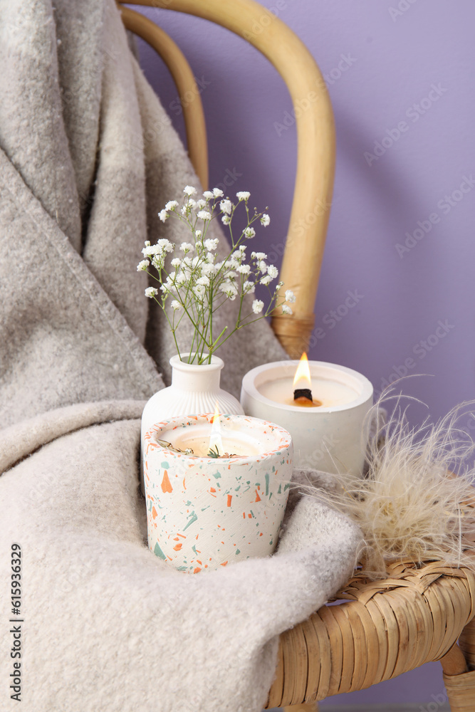 Holders with burning candles, gypsophila flowers and blanket on chair near purple wall in room, clos