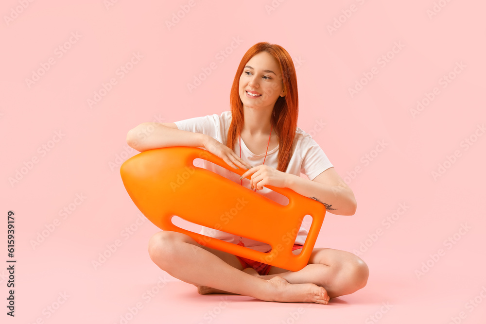 Female lifeguard with rescue buoy sitting on pink background