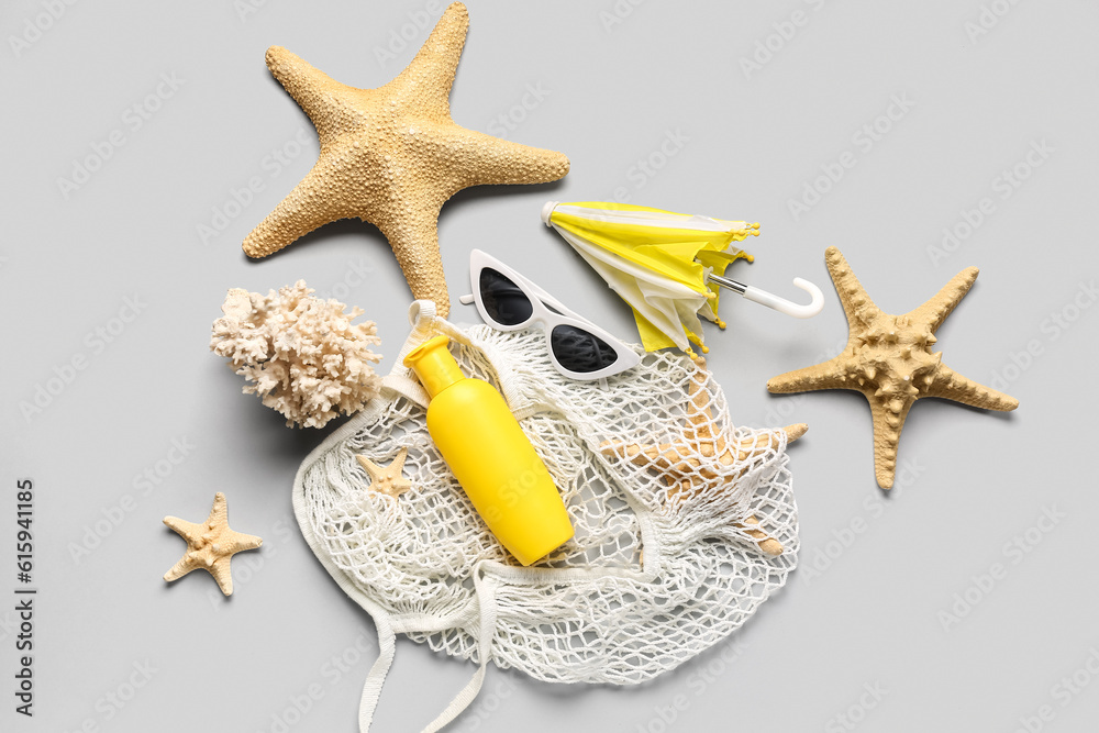 String bag with sunglasses, mini umbrella, coral and bottle of sunscreen cream on grey background