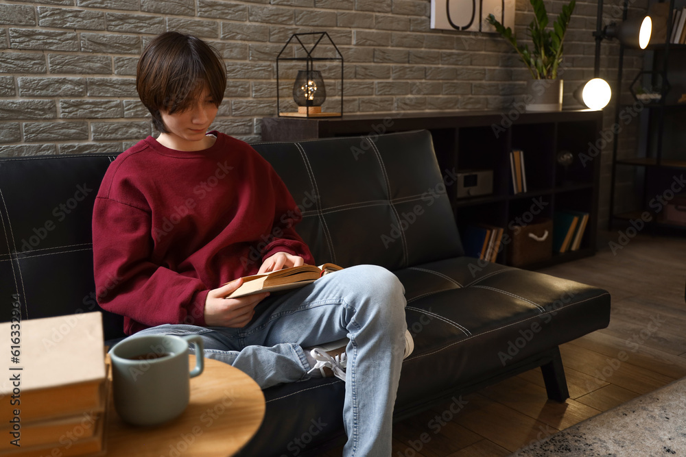 Teenage boy reading book on sofa at home late in evening