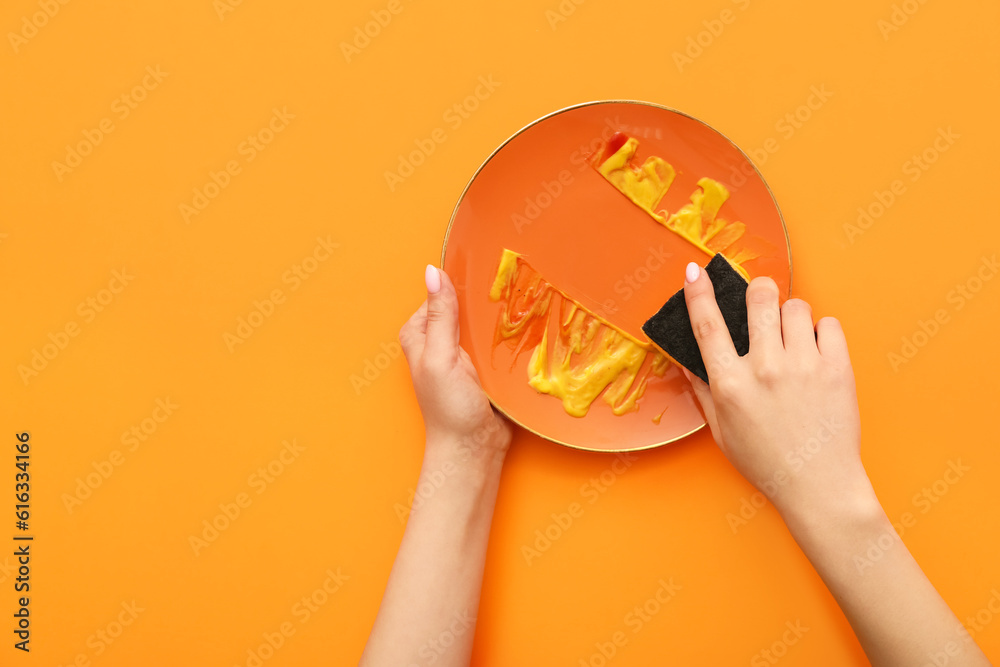 Female hands washing dirty plate with sponge on orange background