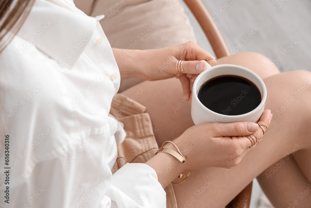 Young woman sitting in armchair and holding cup of delicious coffee