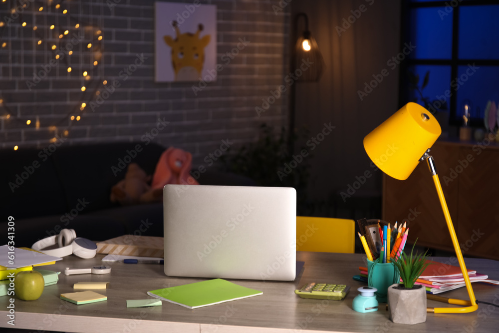 Table with laptop and school stationery in room at night