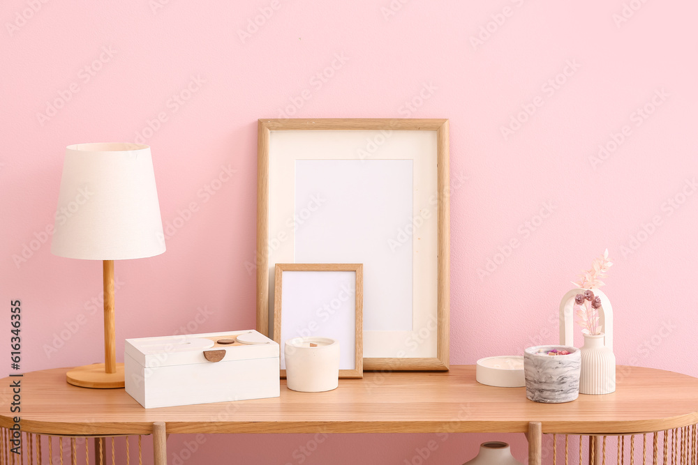 Holders with candles, blank frames, lamp and decor on table near pink wall in room