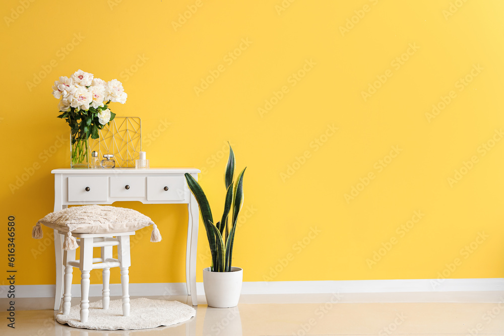 Vase of white peonies with dressing table, chair and houseplant near yellow wall