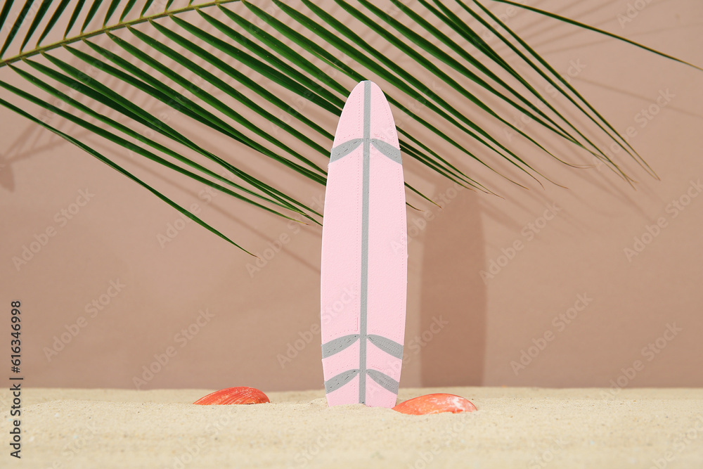 Mini surfboard with seashells and palm leaf on sand against brown background