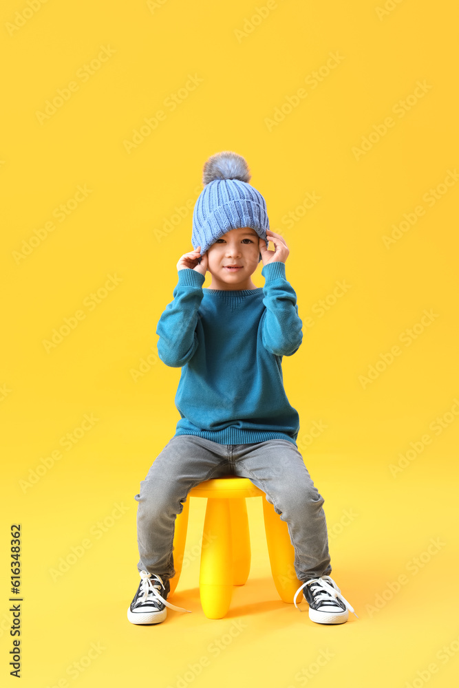 Cute little boy in knitted hat and sweater sitting on yellow background