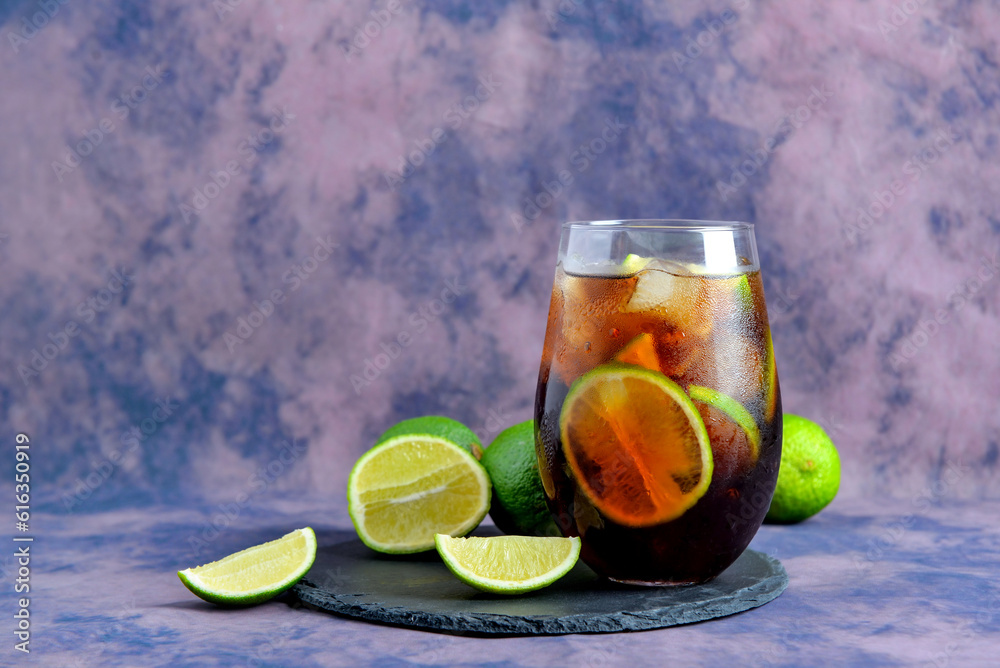 Board with glass of cold Cuba Libre cocktail and limes on purple background