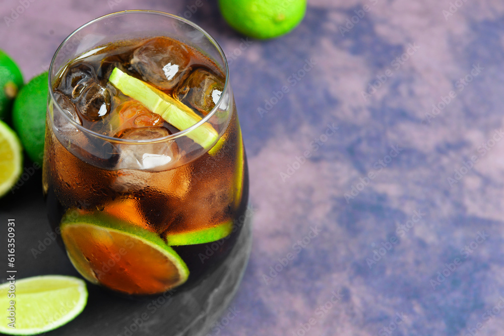 Board with glass of cold Cuba Libre cocktail and limes on purple background