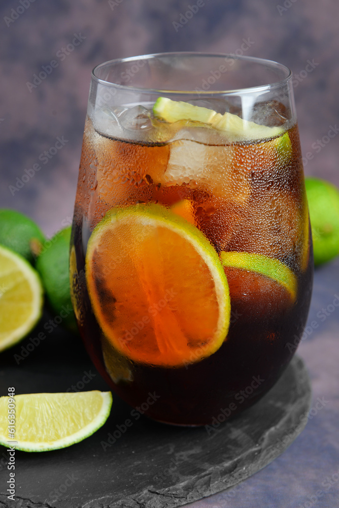 Board with glass of cold Cuba Libre cocktail and limes on purple background