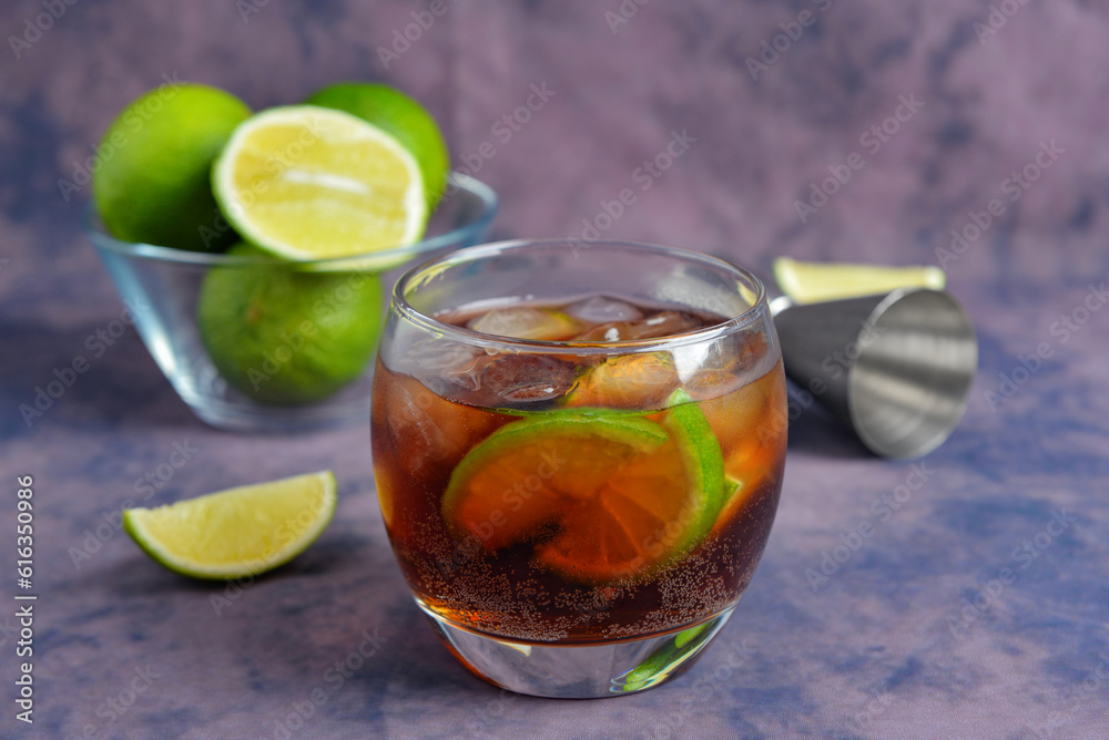 Glass of cold Cuba Libre cocktail and bowl with limes on purple background