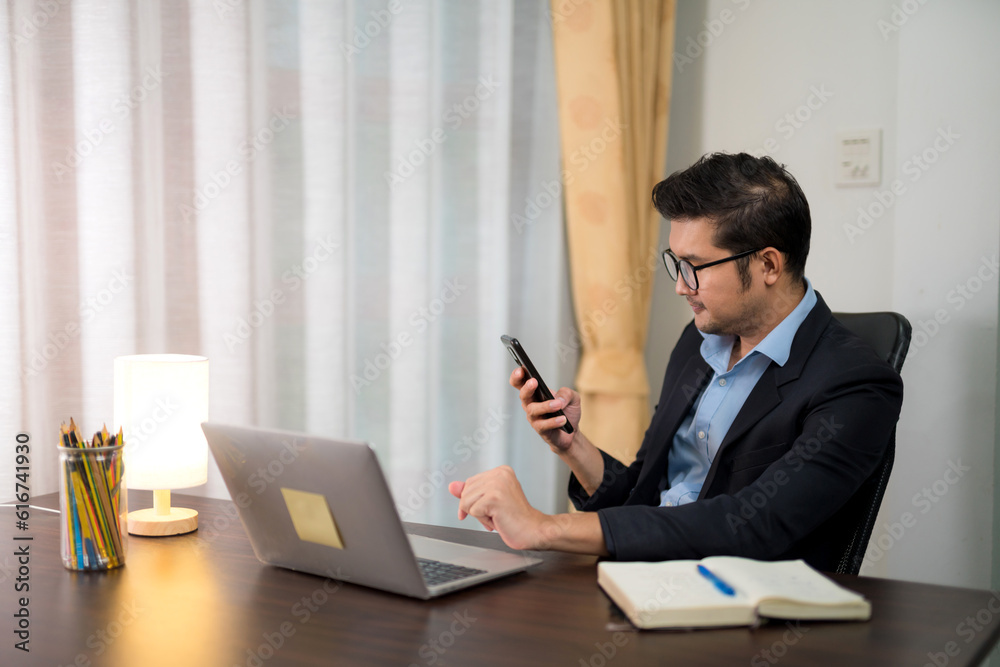 Asian businessman answering a phone call while working on a desk with a laptop at home.