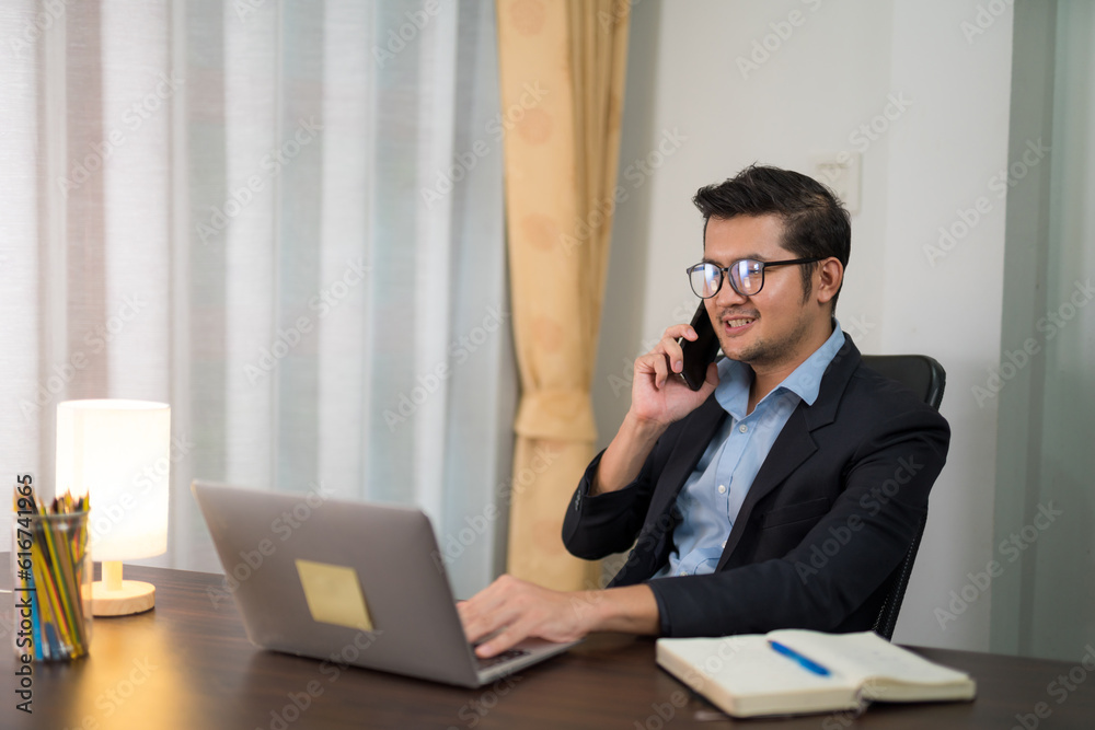 Asian businessman talking on the phone while working on desk with laptop at home.