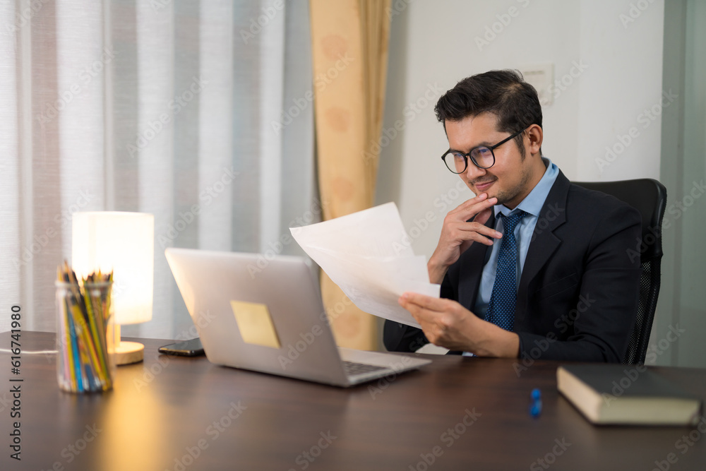 Asian businessman sitting and looking at graph of company sales on desk with laptop in office.