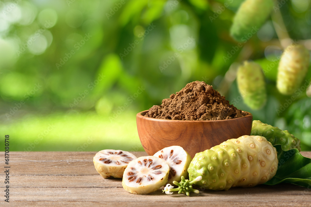 Noni or Morinda Citrifolia powder and fruits on wooden table with blur plant background.