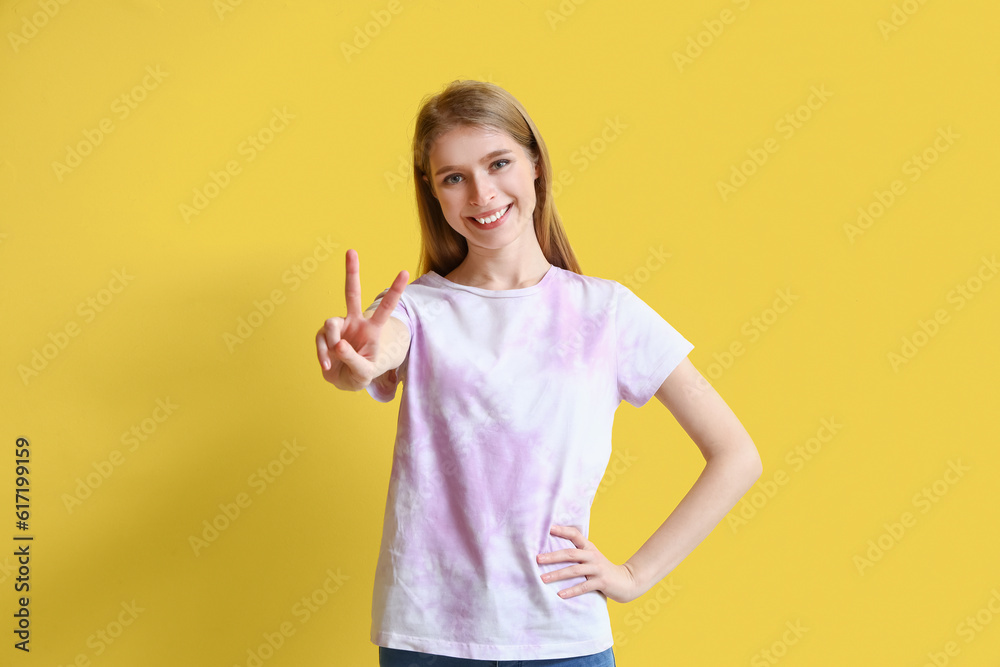 Young woman in tie-dye t-shirt showing victory gesture on yellow background