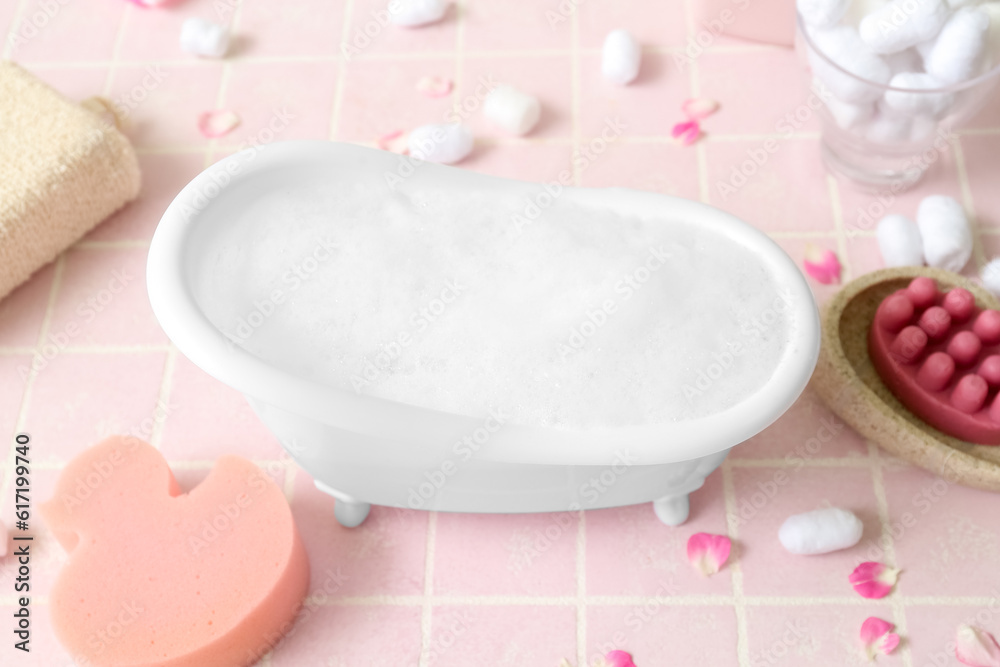 Small bathtub with foam and bath supplies on color tile background