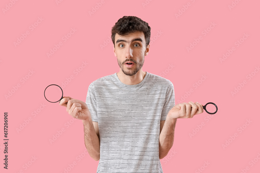 Shocked young man with magnifiers on pink background