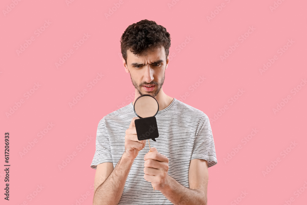 Thoughtful young man with magnifier looking at speech bubble on pink background