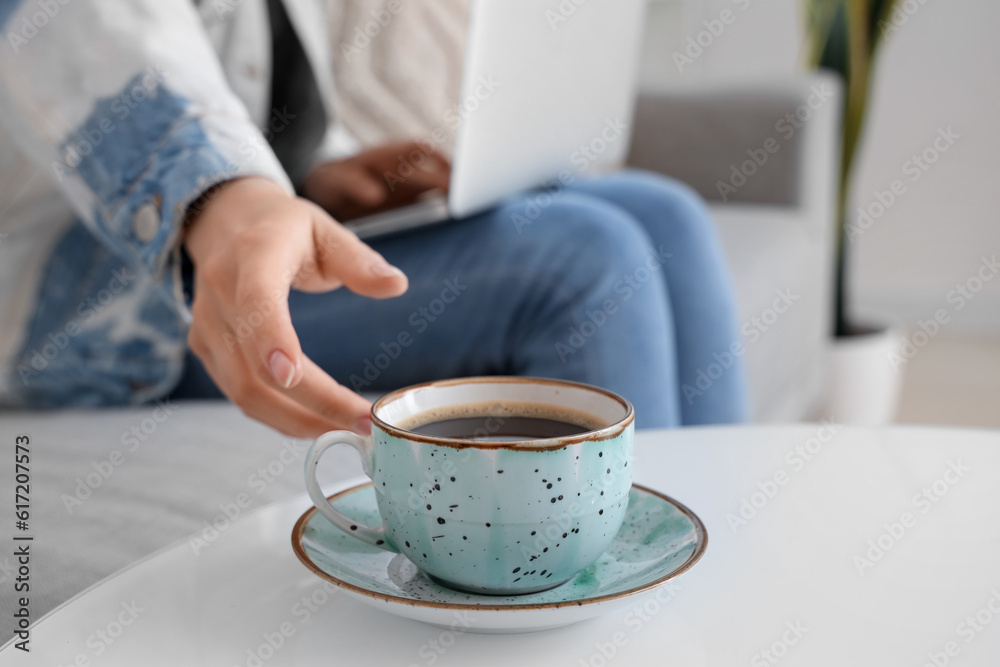 Woman on sofa using laptop and taking cup of coffee from table, closeup