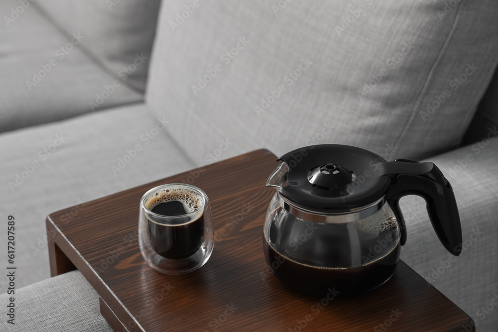 Coffee pot and glass of delicious espresso on table near sofa