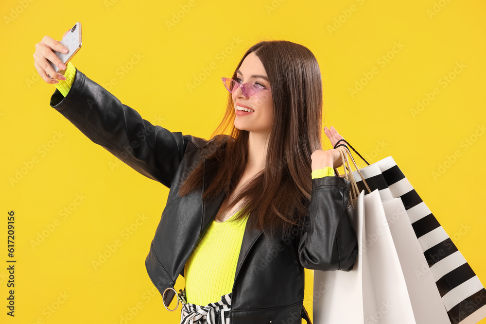 Young woman with shopping bags taking selfie on yellow background