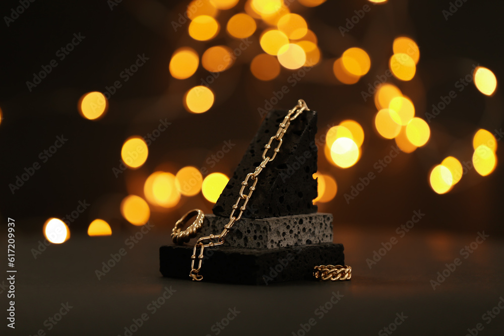 Decorative podium with beautiful chain bracelet and rings on table against blurred lights