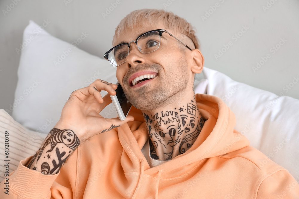 Young tattooed man with eyeglasses talking by mobile phone in bedroom