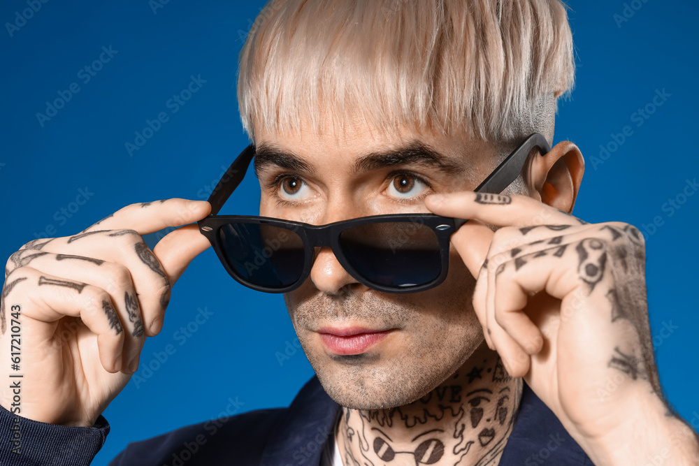 Young tattooed man in sunglasses on blue background, closeup