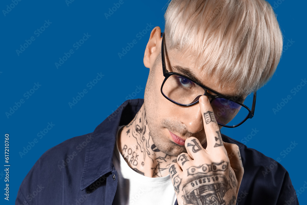 Young tattooed man in eyeglasses on blue background, closeup