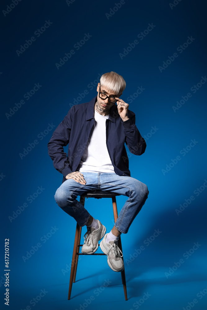 Young tattooed man in eyeglasses sitting on blue background