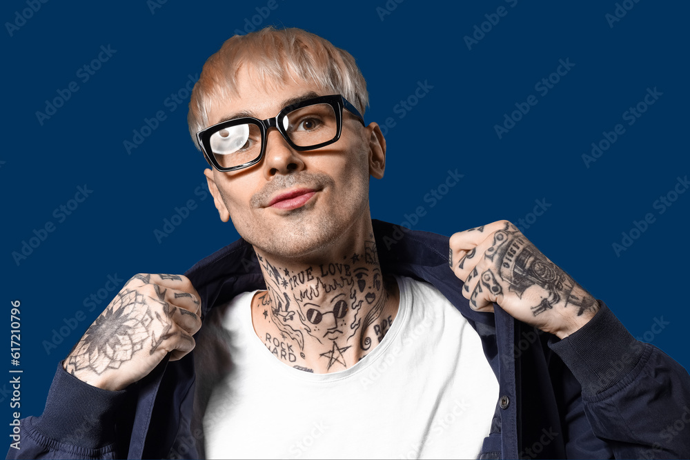 Young tattooed man in eyeglasses on blue background, closeup