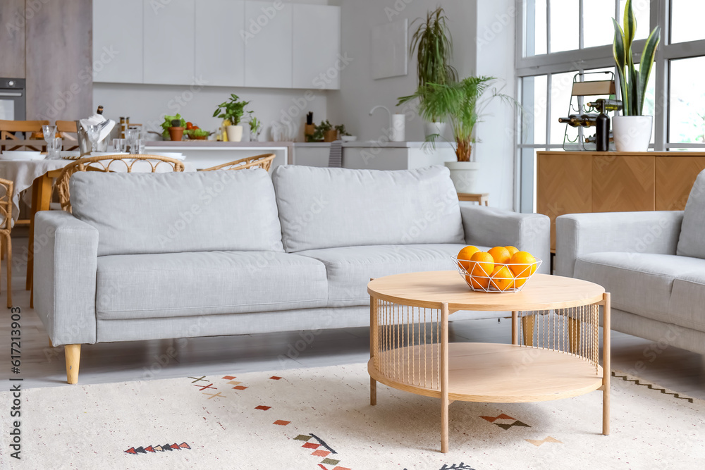 Interior of light open space kitchen with cozy grey sofa and oranges on wooden coffee table