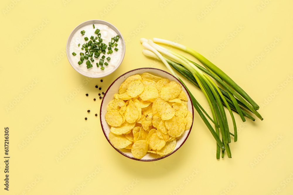 Bowl of tasty sour cream with sliced green onion and potato chips on yellow background