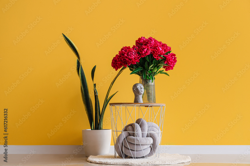 Vase of red peonies with figurine and houseplant near yellow wall