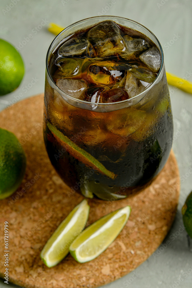 Glass of cold Cuba Libre cocktail and limes on grey background