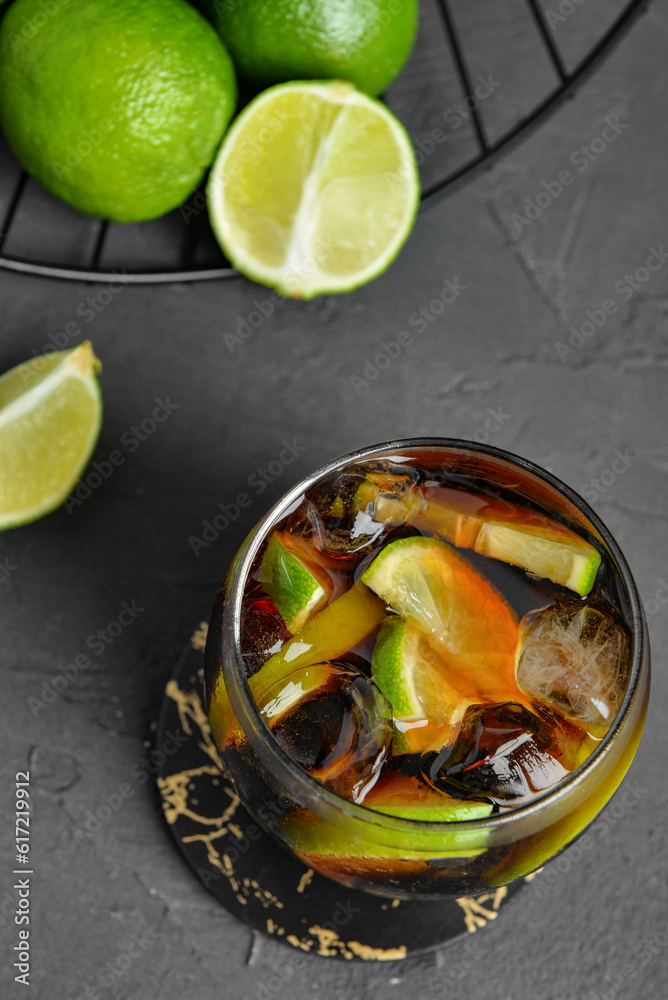 Glass of cold Cuba Libre cocktail and basket with limes on grey table