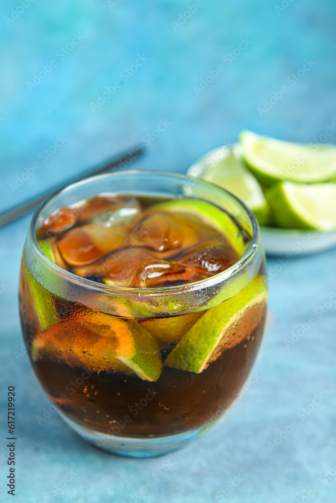 Glass of cold Cuba Libre cocktail and bowl with limes on blue background