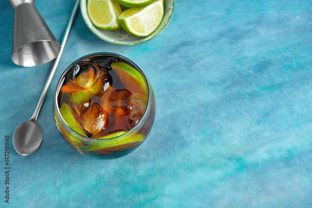 Glass of cold Cuba Libre cocktail and bowl with limes on blue background