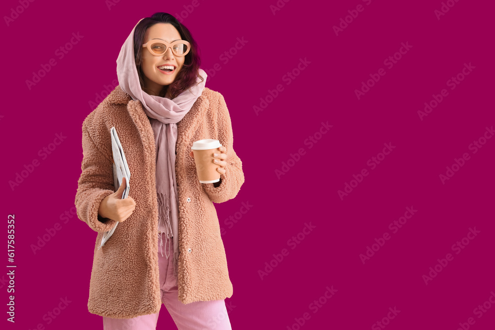 Stylish young woman in teddy coat with cup of coffee and newspaper on pink background