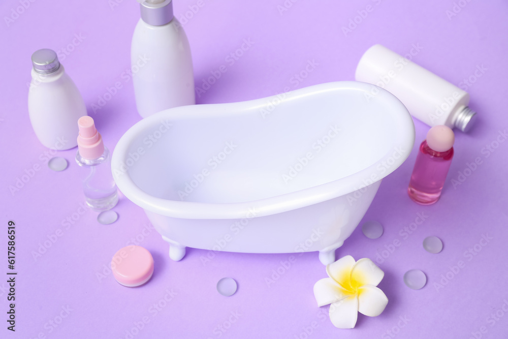 Small bathtub and cosmetic products on lilac background